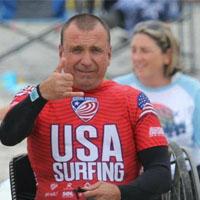 Chaka smiles and wears a red, USA Surfing wet suit while giving a "hang loose" sign.