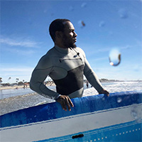 Ty Duckett holds a blue surfboard while staring toward the ocean.