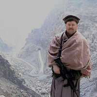 Col Robert W Redding wears tan Afghan clothing while standing on a mountain-lined road between Kabul and Jalalabad.